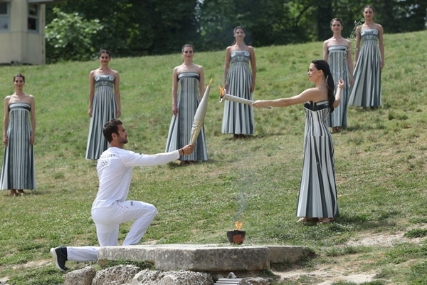 扮演最高女祭司的演員瑪麗·米娜（前右）點燃第一棒火炬手——希臘運動員、東京奧運會男子賽艇項目金牌得主斯特凡諾斯·恩圖斯科斯手中的火炬。新華社記者 趙丁喆 攝